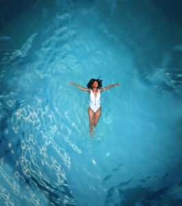 An African American woman in a swimsuit floats peacefully in a clear blue swimming pool.
